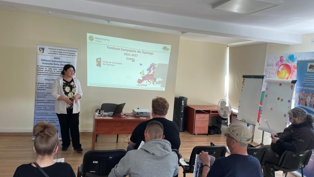 Sala szkoleniowa z kilkoma osobami siedzącymi na krzesłach, skierowanymi w stronę prowadzącej prezentację kobiety. Na ścianie wyświetlona jest prezentacja, a obok znajdują się roll-upy informacyjne oraz tablice z kolorowymi naklejkami.